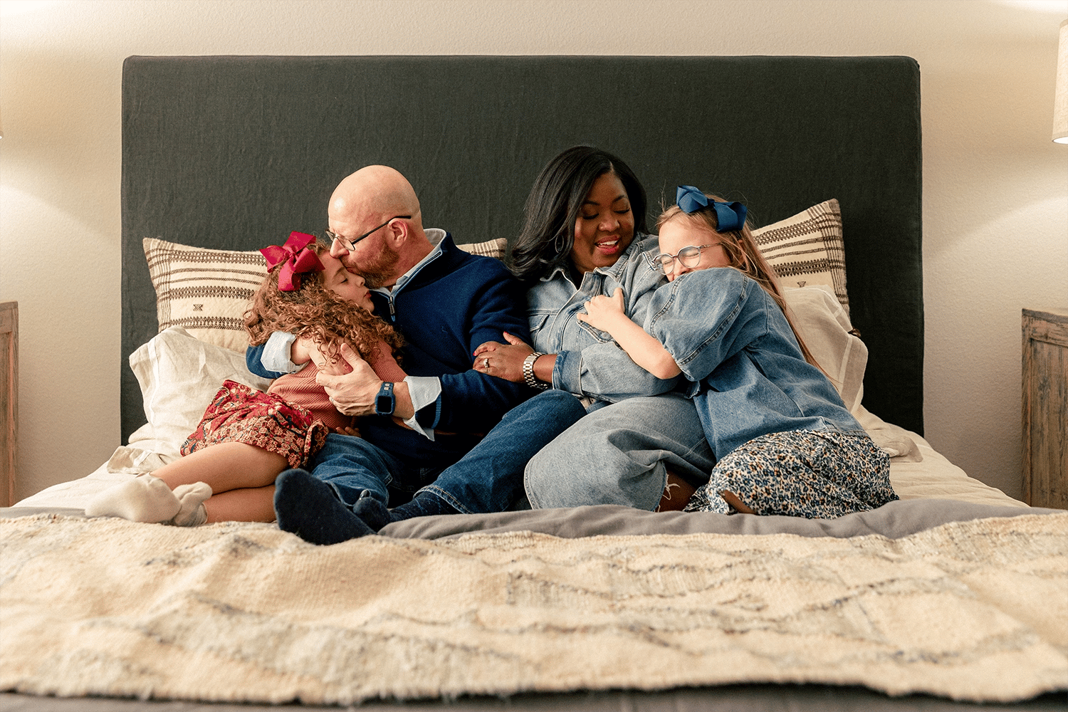 Photo of a family (husband, wife, and two daughters) snuggling together on a bed in the master suite of their Betenbough home. Betenbough homes is one of several home builders in Lubbock Tx.