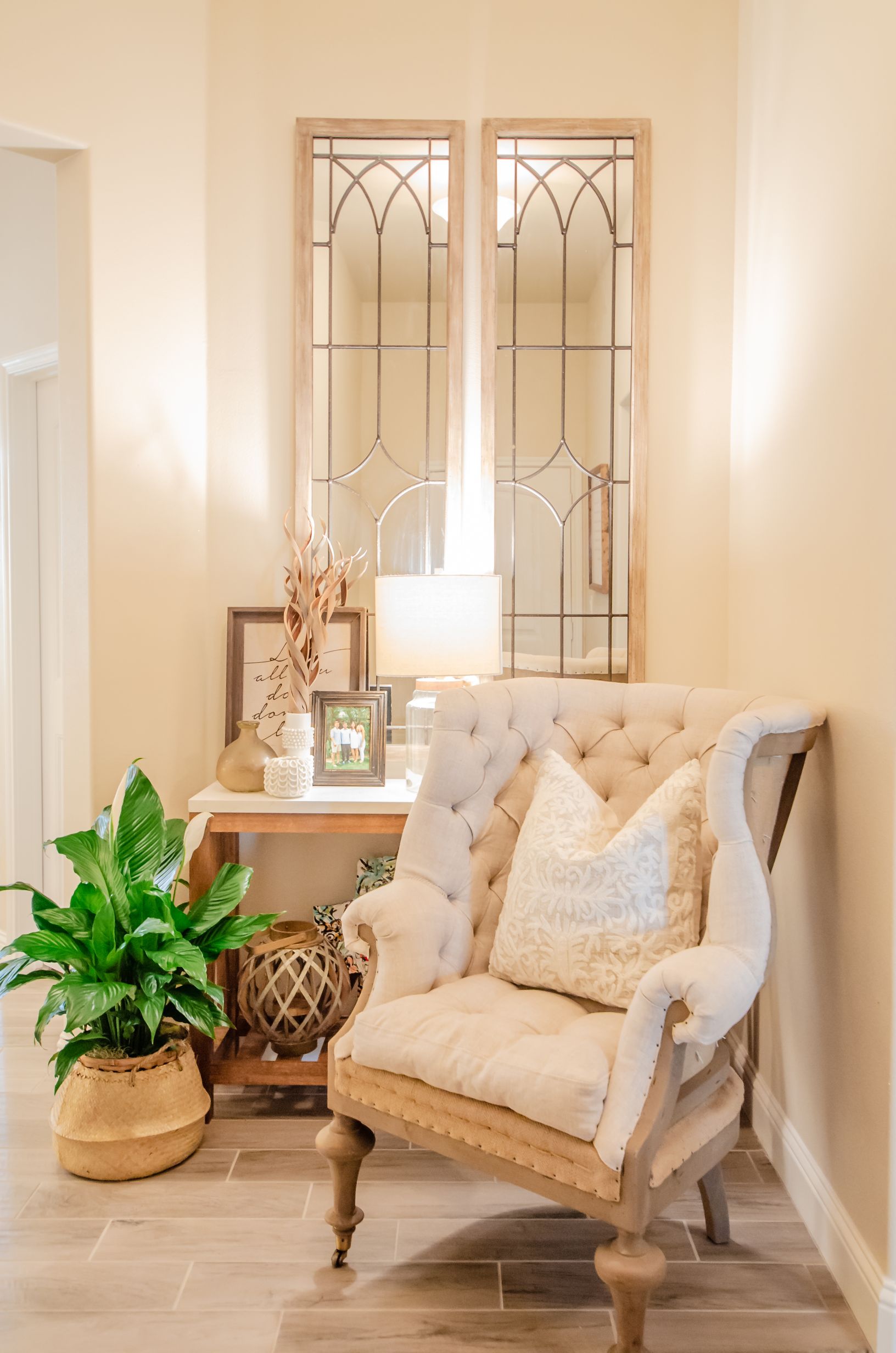 Photo of a decorated home entryway with a decorative mirror, an upholstered chair, a floor plant, and a sofa table decorated with a lamp, photos, and other decor.