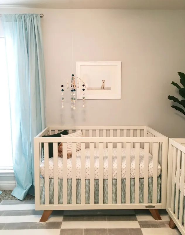 Photo of twin nursery decor. In frame are a curtain, a crib, a mobile, and wall art.