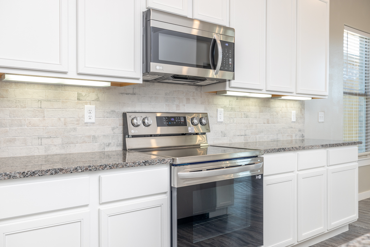 Photo of stainless steel appliances in a Betenbough home's kitchen with white cabinets, a granite countertop, and a tiled backsplash.