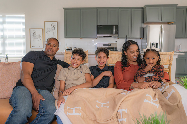 A family of five sits together on a couch in a bright living room, smiling and relaxing under a tan blanket. The parents sit on either side of their three young children, with a modern kitchen visible in the background.