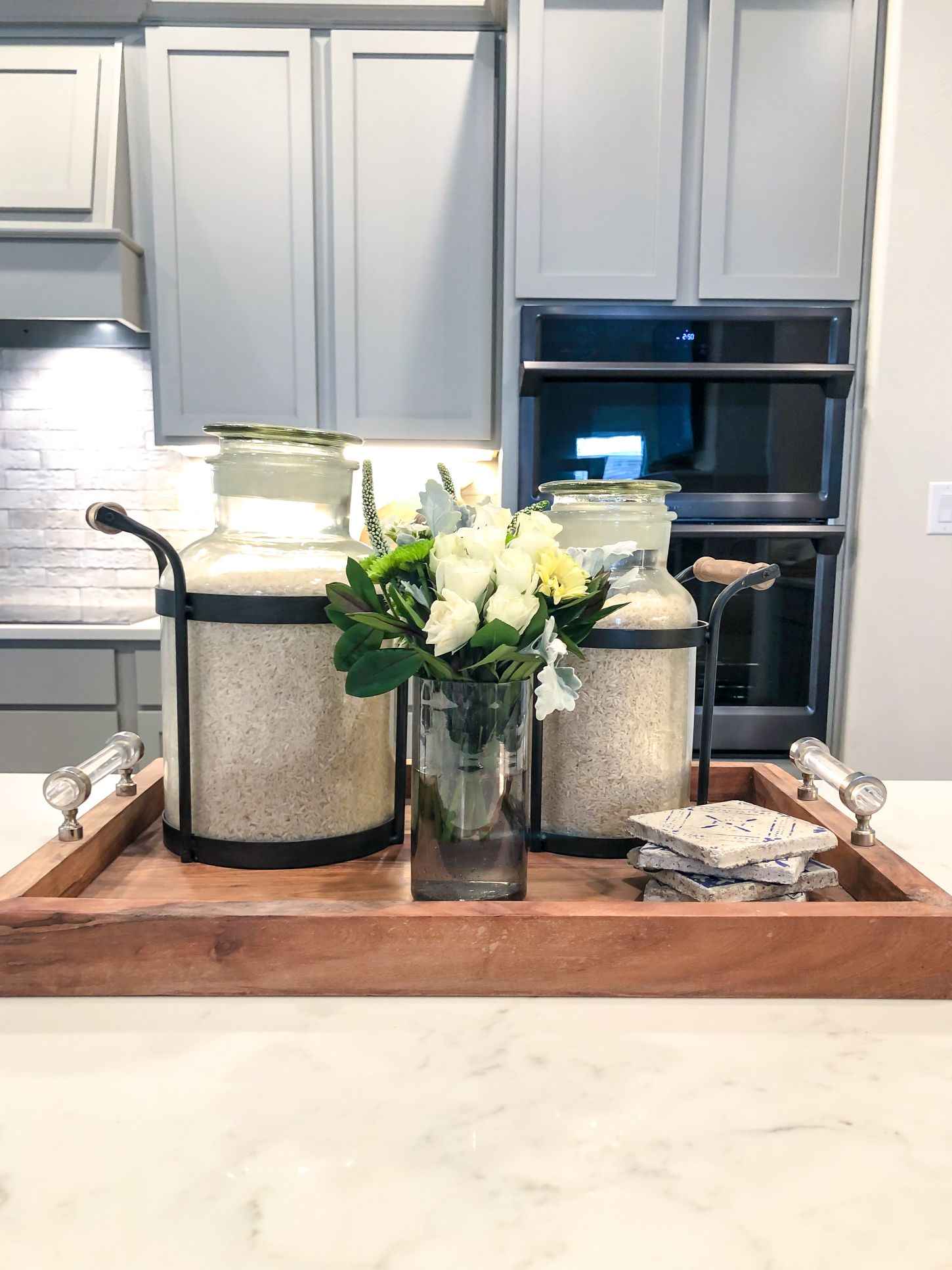 Photo of decorative containers, a vase of fresh flowers, and a set of coasters on a wooden tray sitting on a quartz countertop in a Betenbough Homes kitchen.