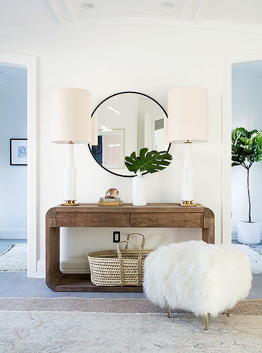 Photo of a decorated home entryway with a round mirror, fur ottoman, and couch table decorated with matching lamps, a plant, books, and a basket.