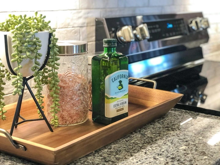 Photo of a plant, container of salt, and a bottle of olive oil, on a wooden tray. The tray sits on a granite countertop next to a flat top stove in a Betenbough home.