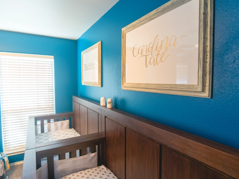 Photo of side-by-side cribs against a blue wall. Framed names