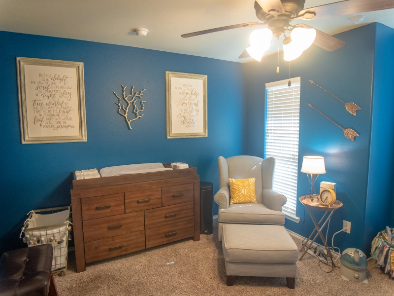 Photo of nursery decor that shows blue walls, a window, a gray chair and ottoman, a side table with a lamp and clock, a changing station, and a laundry hamper.