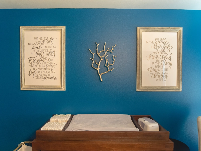 Photo of twin nursery decor shows a blue wall with two framed scriptures and a golden branch are hung above a diaper changing station.