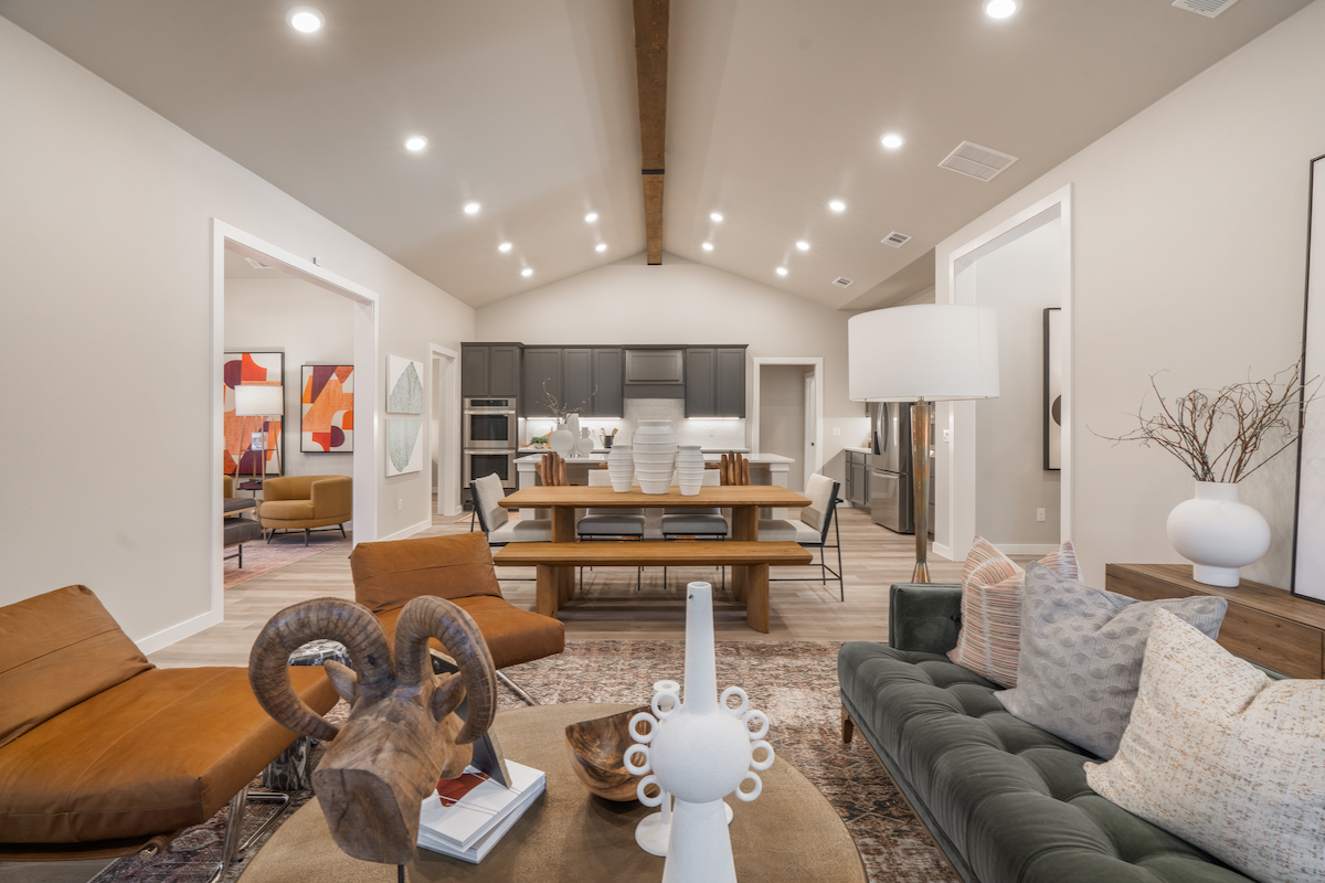 Photo of the Parade Home's living room and kitchen. modern furniture, bright lighting, and a ceiling beam are visible.